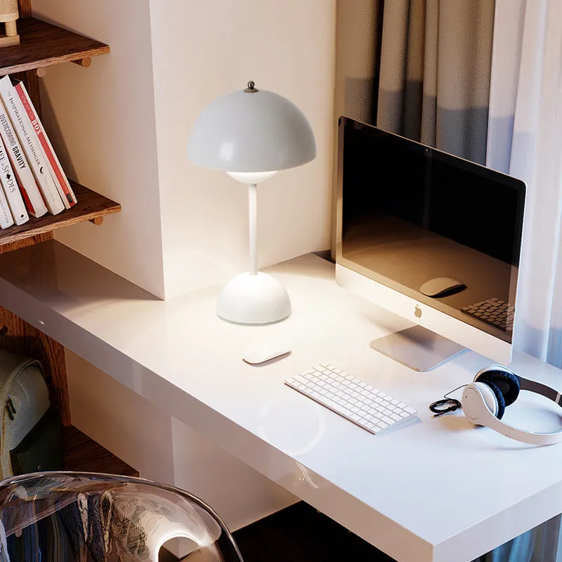 Lampe champignon blanche allumée sur un bureau blanc avec écran, clavier, souris et casque audio, près d'une étagère de livres. 7