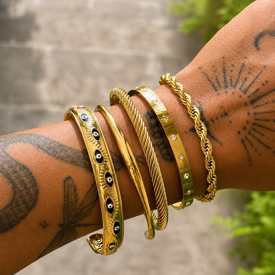 Poignet tatoué portant le jonc doré lisse, empilé avec plusieurs autres bracelets dorés torsadés, ornés d'yeux et de petites pierres. 3
