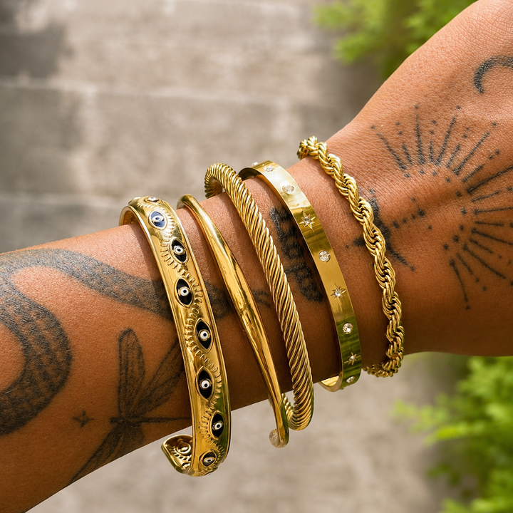 Poignet tatoué portant le jonc doré lisse, empilé avec plusieurs autres bracelets dorés torsadés, ornés d'yeux et de petites pierres. 3