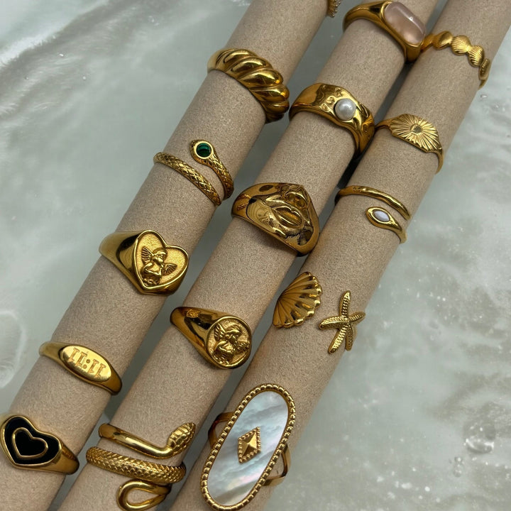 Collection de bagues dorées présentées sur des tubes beiges, dont la bague cœur avec ange, des bagues serpent, coquillage et perle. 5