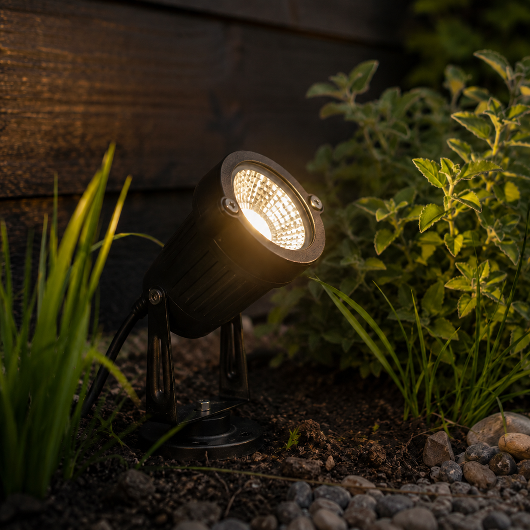 Vue rapprochée du projecteur noir allumé au sol, lumière chaude, avec son câble, son piquet et sa base, parmi des plantes et galets. 6