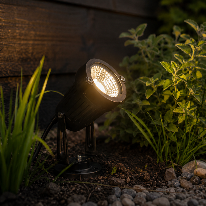 Vue rapprochée du projecteur noir allumé au sol, lumière chaude, avec son câble, son piquet et sa base, parmi des plantes et galets. 6
