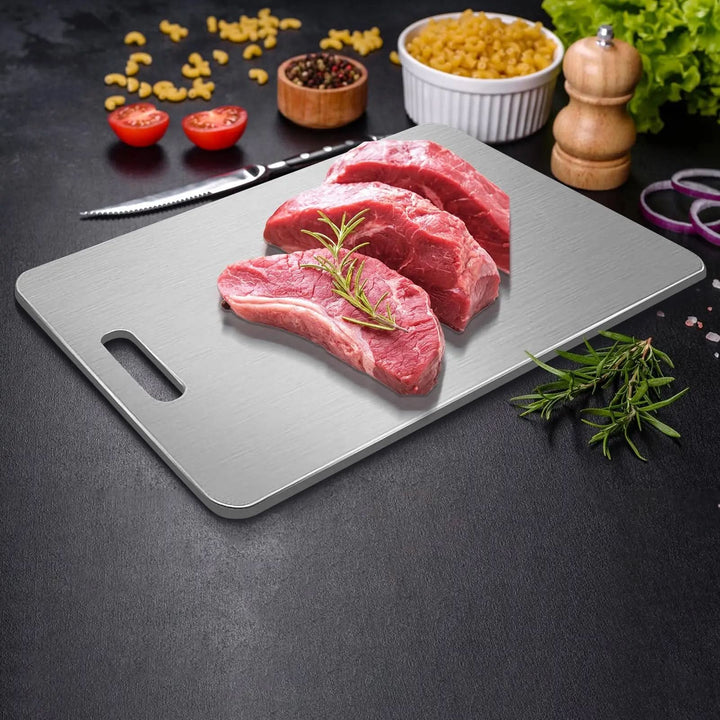 Trois morceaux de viande crue garnis de romarin sur la planche en inox, entourée d'épices, de pâtes, de tomates et d'un couteau. 4