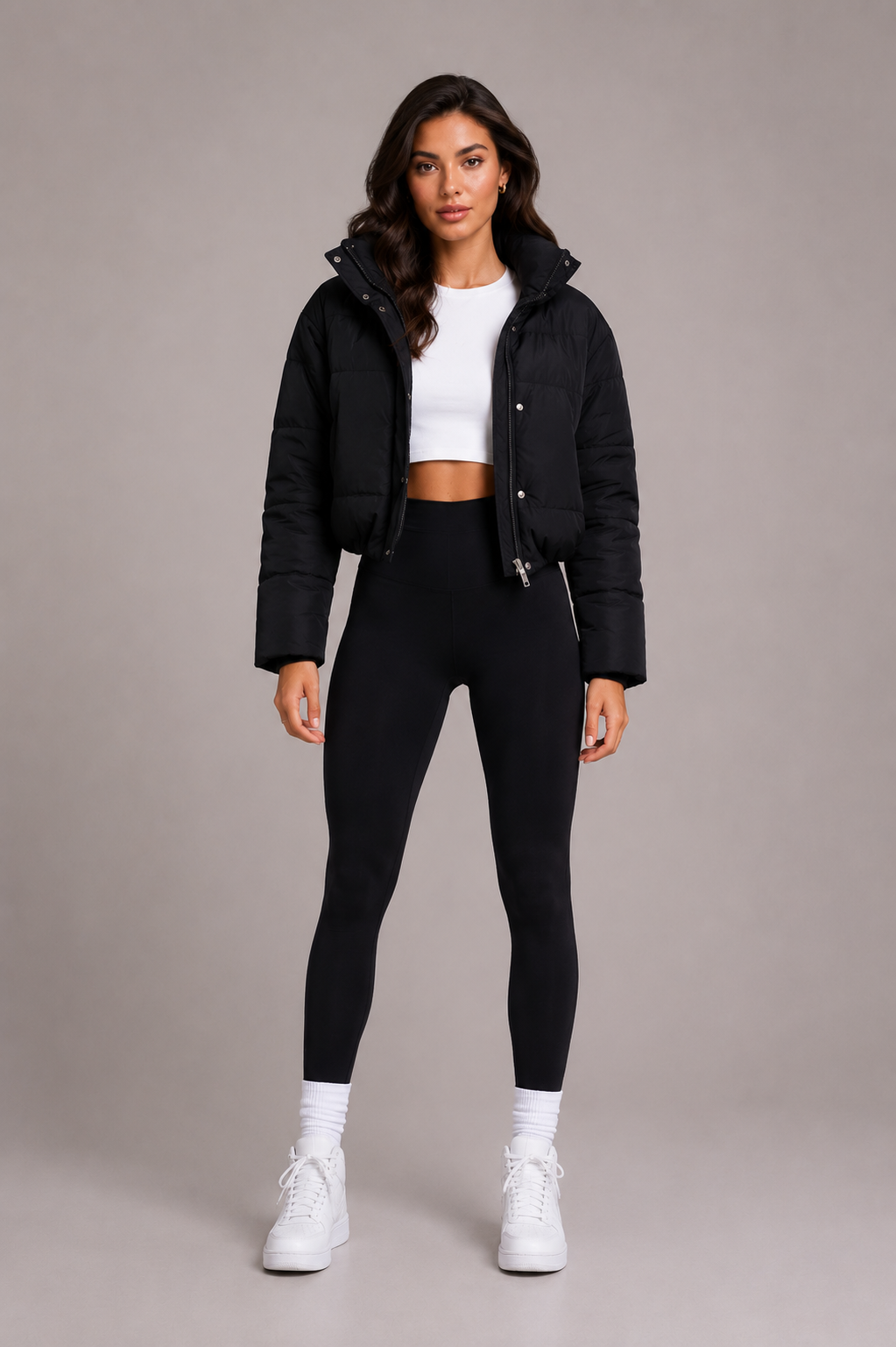 Femme en pied de face, doudoune noire courte ouverte sur un crop top blanc, legging noir et baskets blanches. 2