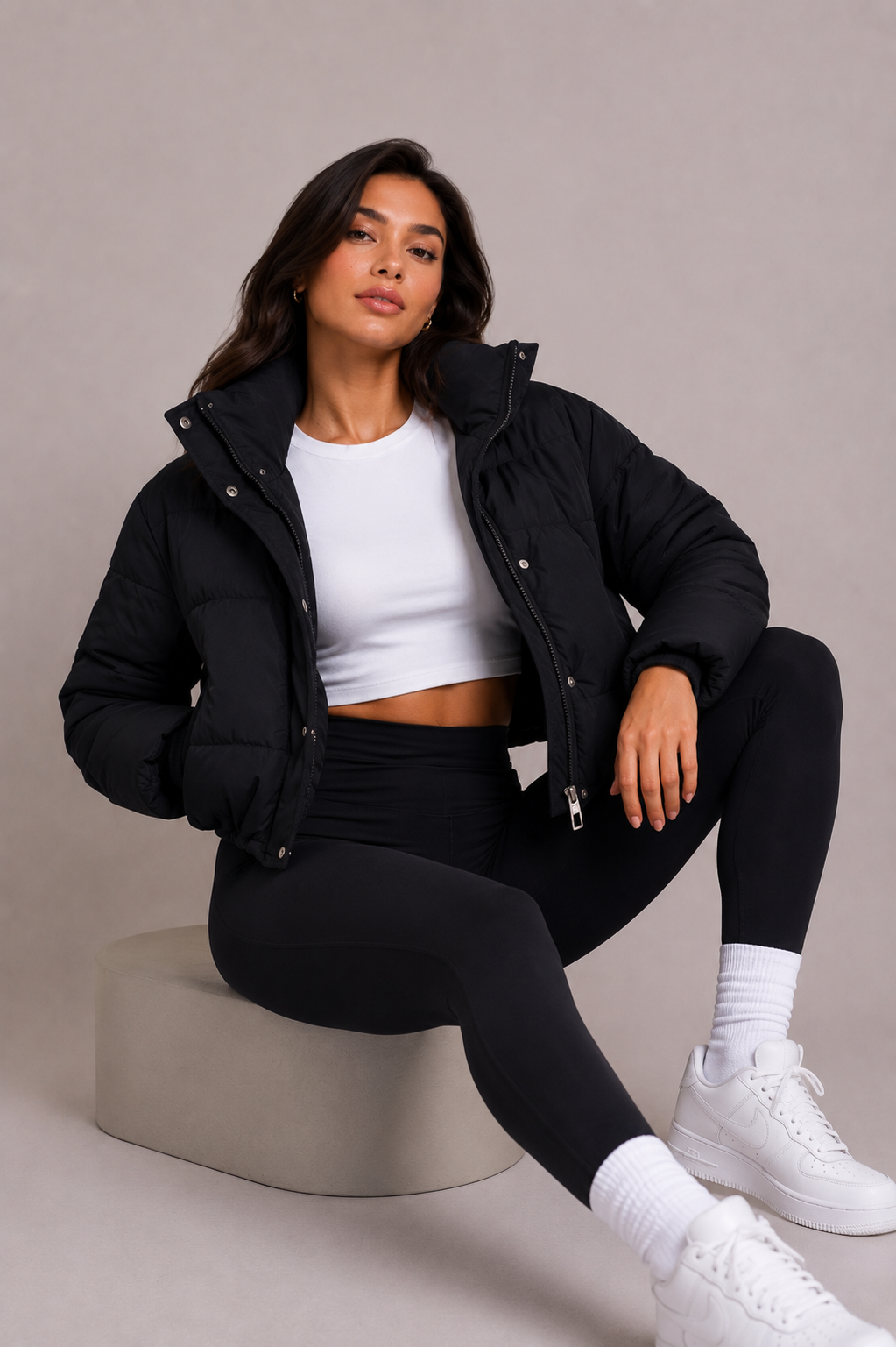 Femme assise sur un tabouret beige, doudoune noire ouverte sur un haut blanc, posant en regardant l'objectif. 6