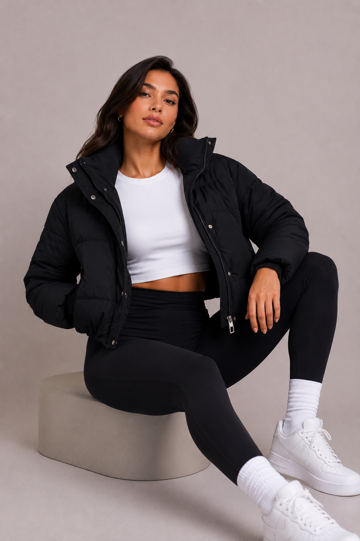 Femme assise sur un tabouret beige, doudoune noire ouverte sur un haut blanc, posant en regardant l'objectif. 6