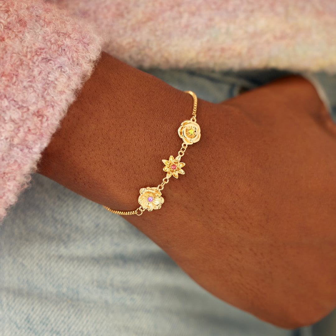 Poignet d'une femme portant le bracelet doré à trois fleurs, manche rose duveteuse et jean clair en arrière-plan. 9