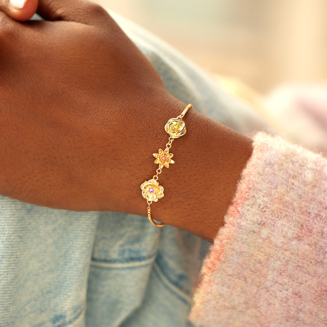 Bracelet doré à trois fleurs porté au poignet, main posée sur un jean clair, avec une manche en maille rose pastel. 10