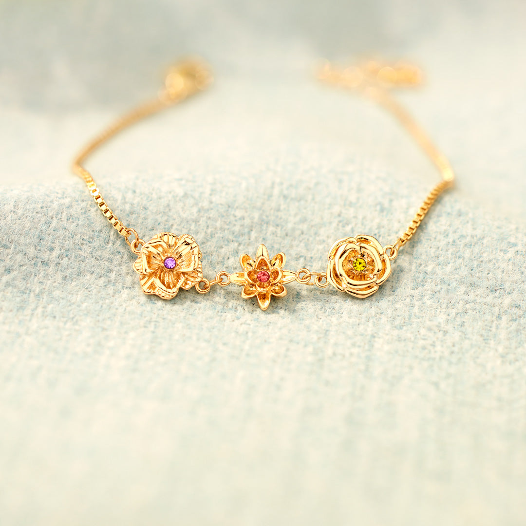 Bracelet doré aux trois fleurs et pierres colorées, posé sur une étoffe bleu clair en laine, avec un arrière-plan flou. 15