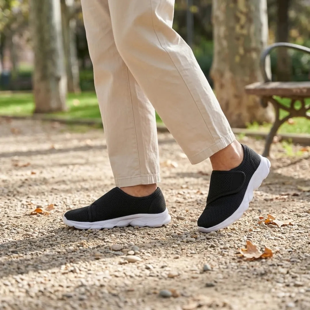 Pieds d'un marcheur chaussés de baskets noires à semelle blanche et sangle, pantalon beige, sur un chemin de parc. 20