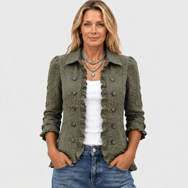 Vue de face, la veste kaki à fleurs brodées portée ouverte sur un top blanc, avec un collier turquoise et les mains dans les poches. 2