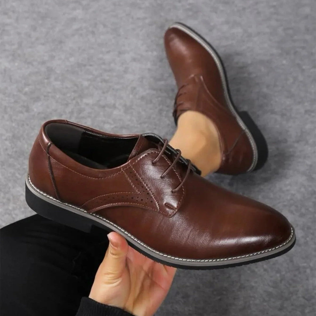 Chaussure marron foncé tenue à la main, vue de dessus, avec détail de perforations et coutures apparentes sur un fond gris. 2