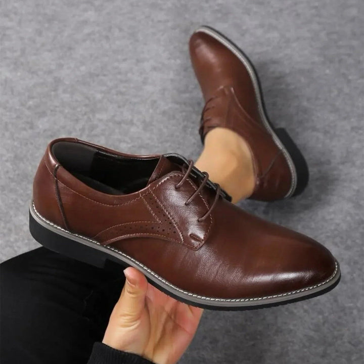 Chaussure marron foncé tenue à la main, vue de dessus, avec détail de perforations et coutures apparentes sur un fond gris. 2