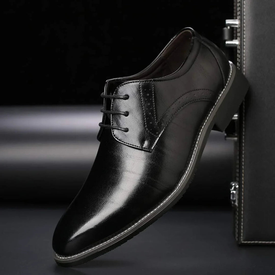 Chaussure noire en cuir lisse et brillant, présentée de profil contre une mallette sombre, avec perforations sur le côté. 3