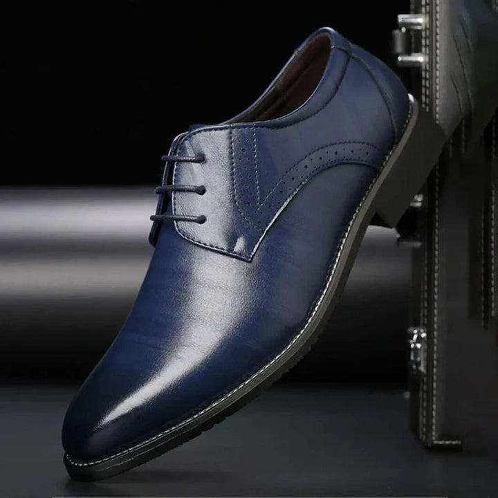 Chaussure bleu marine en cuir lustré présentée de profil contre une mallette noire, avec trois œillets et perforations décoratives. 5