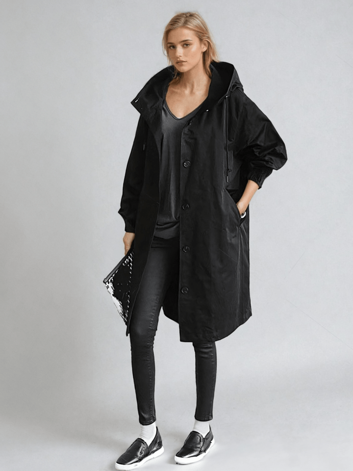 Femme de face en trench noir à capuche, porté ouvert sur un top gris foncé, jean noir et baskets, pochette à la main. 2
