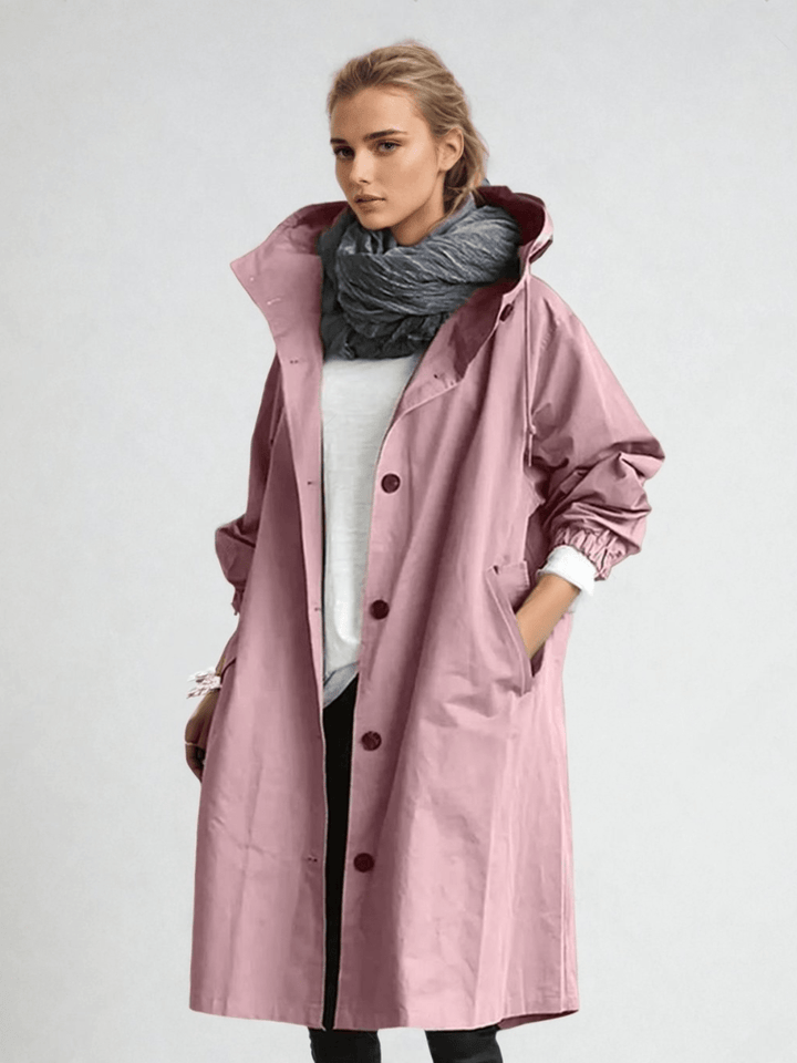 Femme portant le trench à capuche rose poudré boutonné, écharpe grise, haut blanc et pantalon noir, main dans la poche. 6