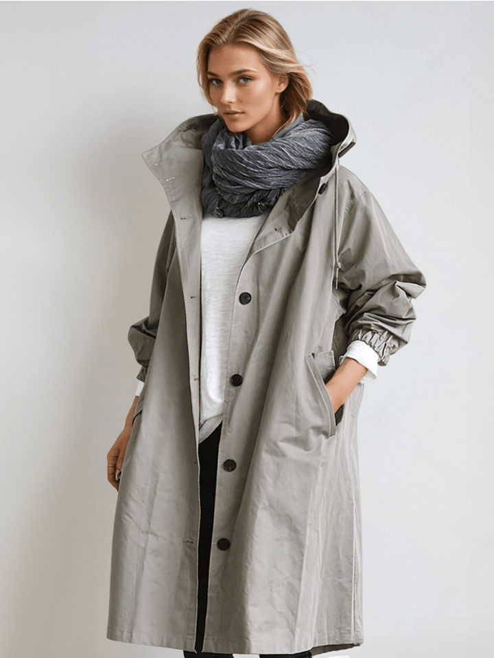 Femme portant le trench à capuche gris clair boutonné, avec écharpe grise et t-shirt blanc, une main dans la poche. 1