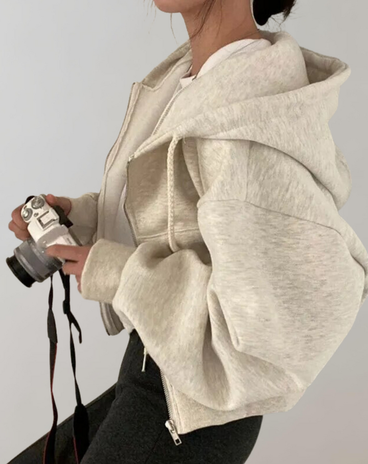 Vue de profil du sweat gris clair chiné porté par une femme tenant un appareil photo, capuche et manche ample visibles. 2