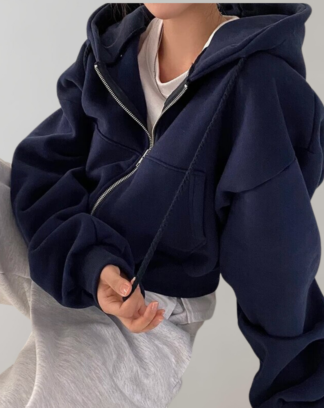 Sweat bleu marine zippé porté avec un t-shirt blanc et un pantalon gris, main tenant un cordon de capuche. 3