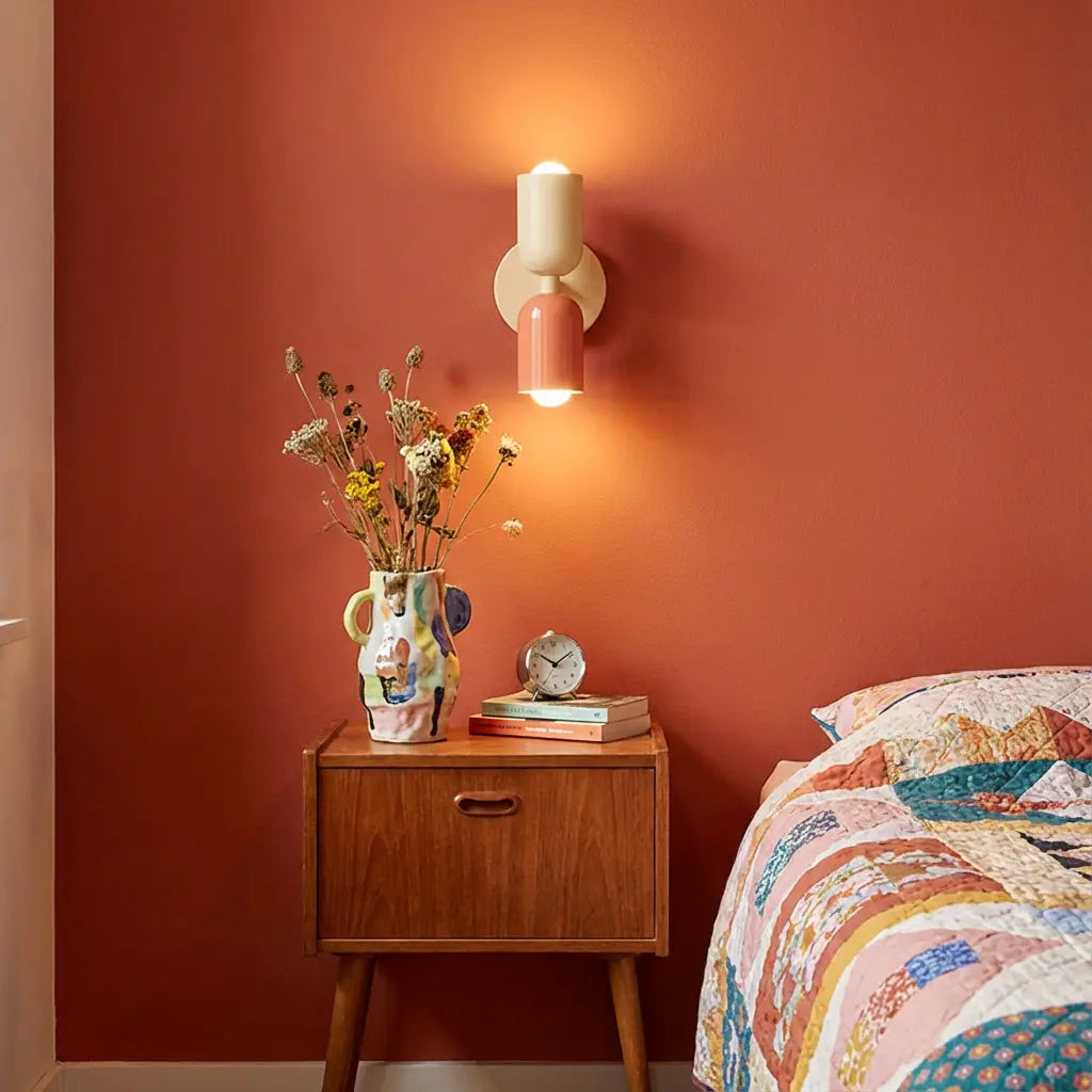 Applique allumée, tête crème et tête corail, sur un mur terracotta au-dessus d'une table de chevet en bois avec vase fleuri. 1