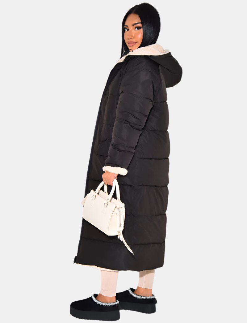 Femme de profil portant le manteau matelassé noir fermé, capuche baissée, col et poignet en fourrure beige, longueur jusqu'aux chevilles. 6