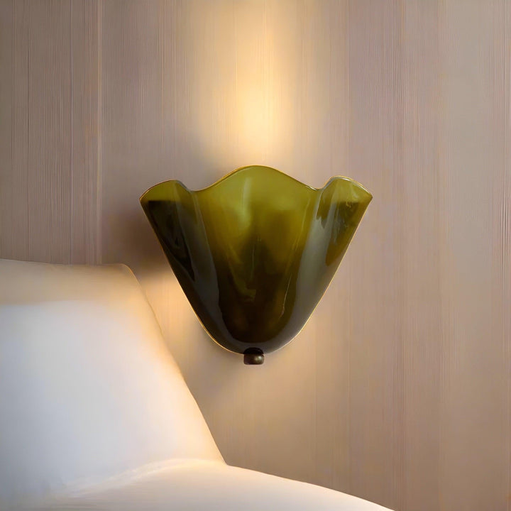 Gros plan d'une applique en verre vert olive à bord ondulé, allumée, contre un mur strié près d'un coussin. 2