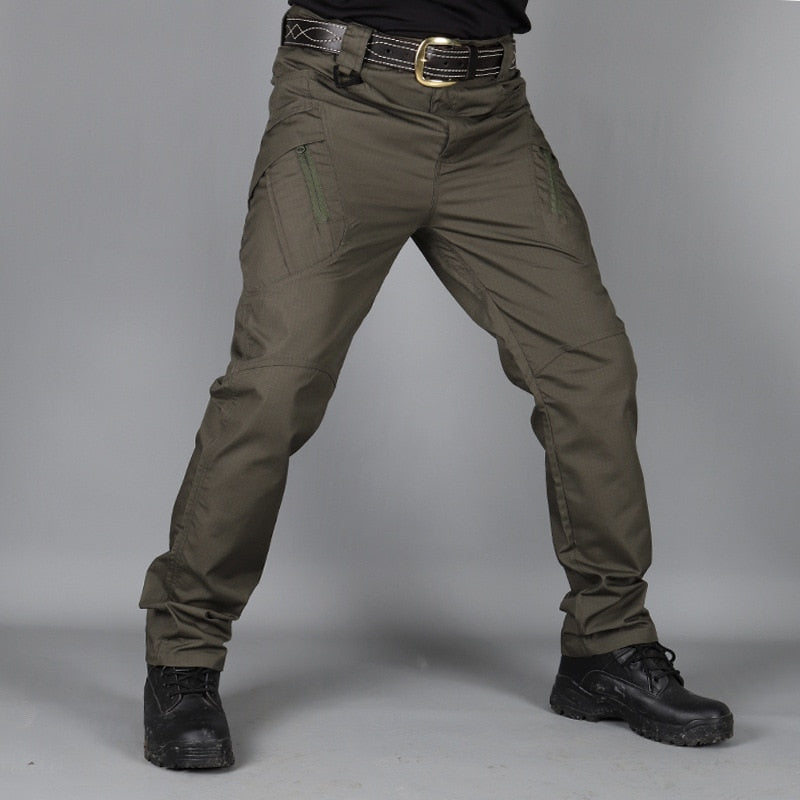 Pantalon vert olive porté de face par un modèle avec ceinture en cuir, poches cargo zippées et chaussures montantes noires sur fond gris. 4