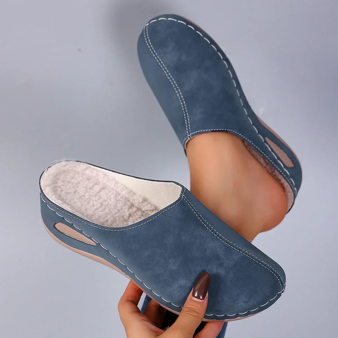 Paire de chaussons bleus tenue en main, doublure en polaire beige visible, coutures apparentes et découpe latérale sur la semelle compensée. 1