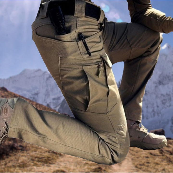 Pantalon cargo beige porté en position agenouillée devant des montagnes enneigées, avec poches à fermeture éclair et poche de cuisse bien visibles. 1