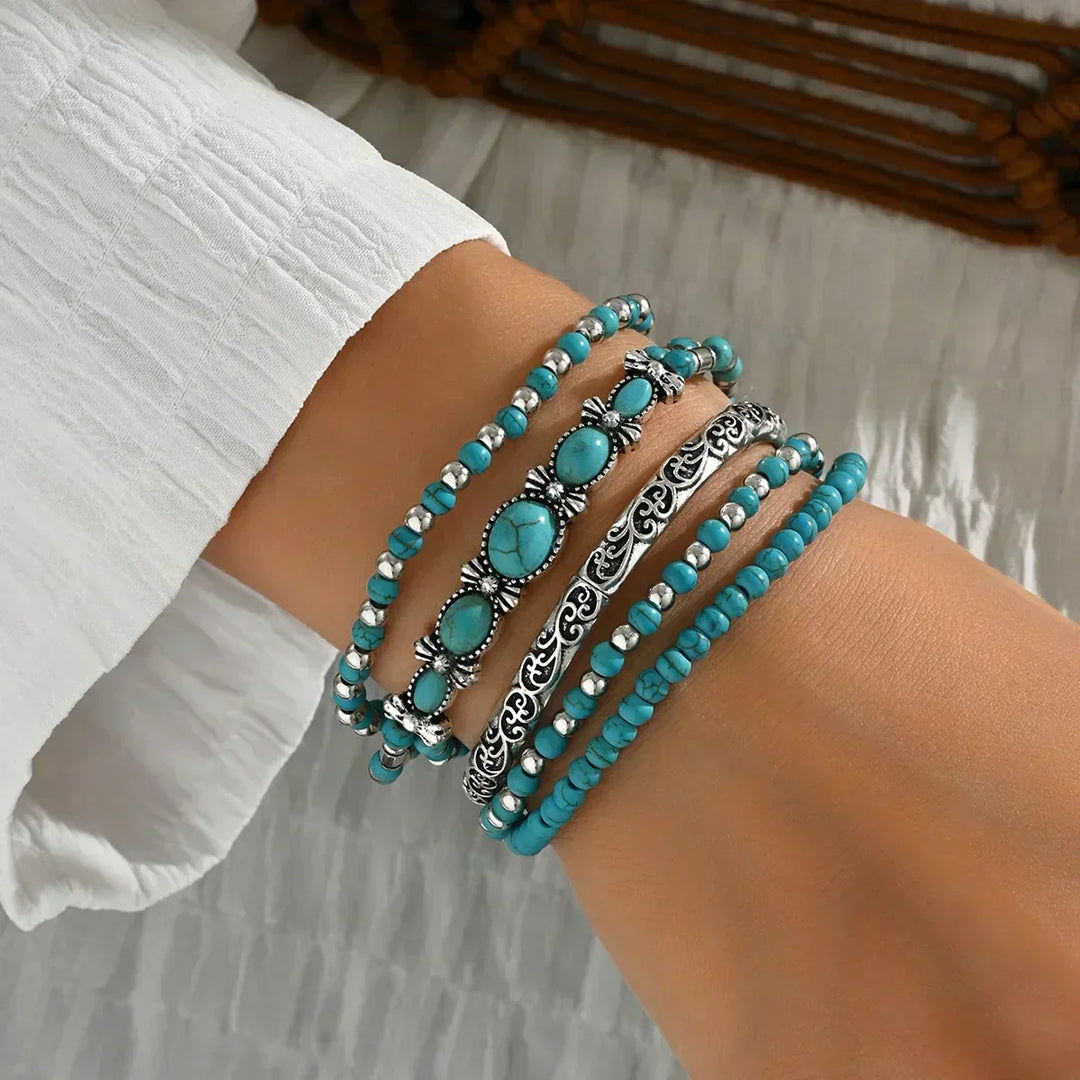 Poignet portant les cinq bracelets empilés, perles turquoise, perles argentées et bracelet ciselé, devant une manche blanche en coton froissé. 1