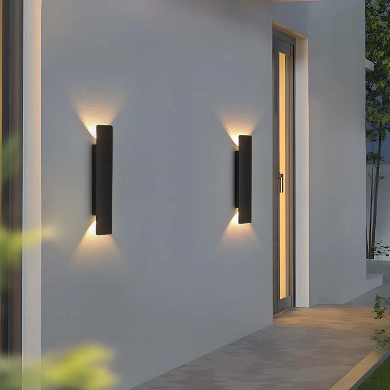 Deux appliques noires fixées sur un mur gris près d'une porte vitrée éclairée, projetant des cônes de lumière chaude vers le haut et le bas. 6