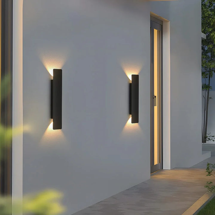 Deux appliques noires fixées sur un mur gris près d'une porte vitrée éclairée, projetant des cônes de lumière chaude vers le haut et le bas. 6