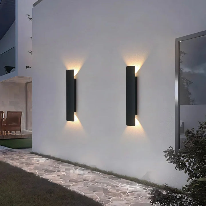 Deux appliques noires rectangulaires sur un mur extérieur blanc le long d'une allée dallée, projetant des faisceaux lumineux chauds vers le haut et le bas. 2