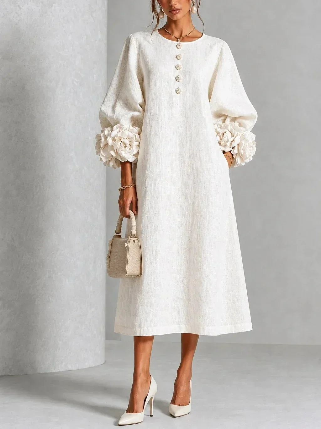 Robe blanc crème portée de face, manches bouffantes ornées de grosses fleurs en relief, boutons fleuris, sac à main beige et escarpins. 1