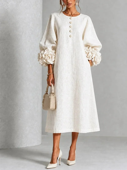 Robe blanc crème portée de face, manches bouffantes ornées de grosses fleurs en relief, boutons fleuris, sac à main beige et escarpins. 1