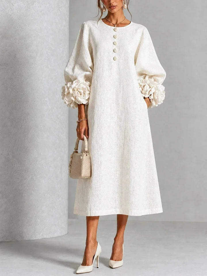 Robe blanc crème portée de face, manches bouffantes ornées de grosses fleurs en relief, boutons fleuris, sac à main beige et escarpins. 1