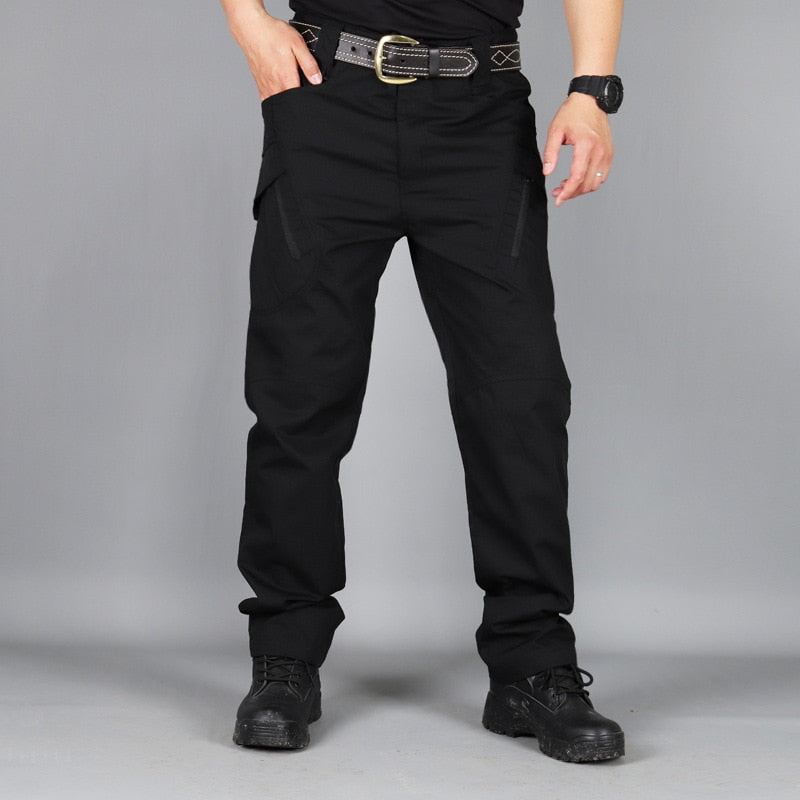 Pantalon noir porté de face, une main dans la poche, avec ceinture à boucle dorée et chaussures montantes noires sur fond gris. 5
