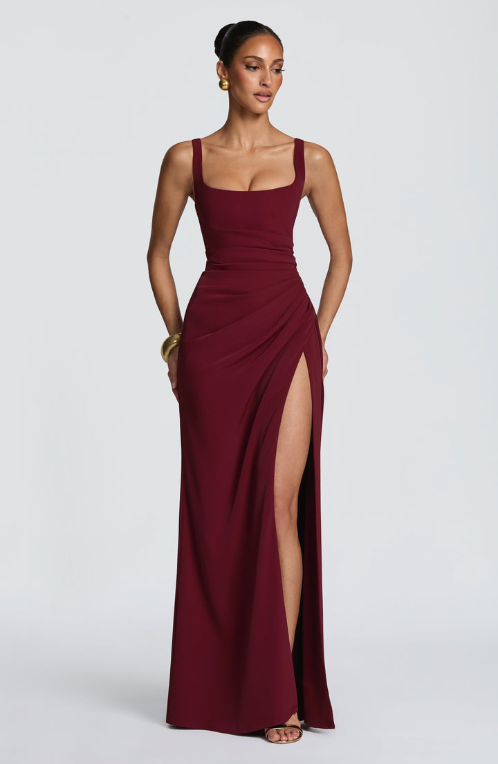 Robe longue bordeaux portée de face, encolure carrée, taille drapée et fente haute, avec bijoux dorés. 17