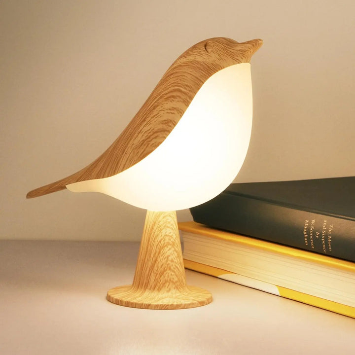 Lampe oiseau allumée, au corps blanc lumineux et à la tête effet bois clair, posée près de livres 1