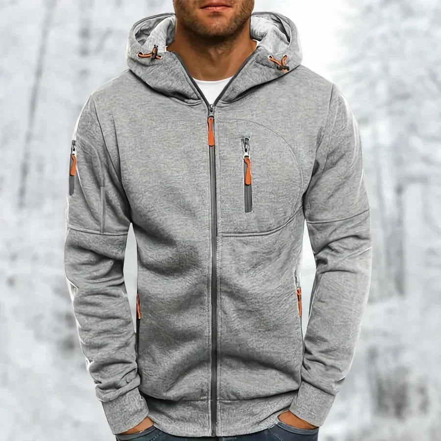 Sweat à capuche zippé gris clair chiné porté par un modèle, tissu jacquard texturé, zips orange, fond enneigé. 8
