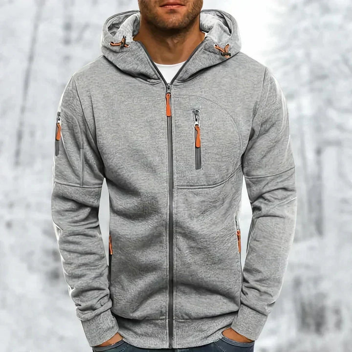 Sweat à capuche zippé gris clair chiné porté par un modèle, tissu jacquard texturé, zips orange, fond enneigé. 8