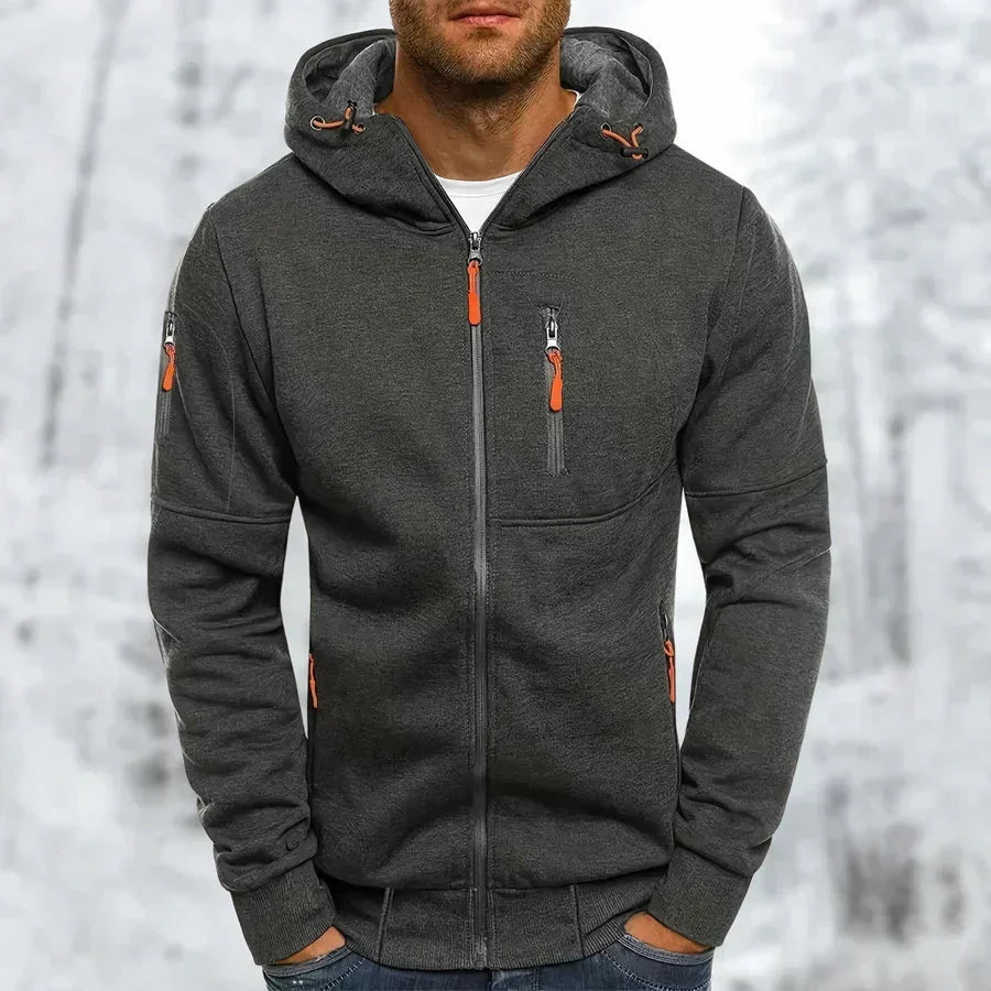 Sweat à capuche zippé gris anthracite porté par un modèle, zips orange, capuche doublée et poignets côtelés, fond enneigé. 9