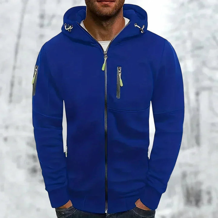 Homme portant le sweat zippé bleu roi à capuche, zips vert citron et poche poitrine, sur fond de forêt enneigée. 1