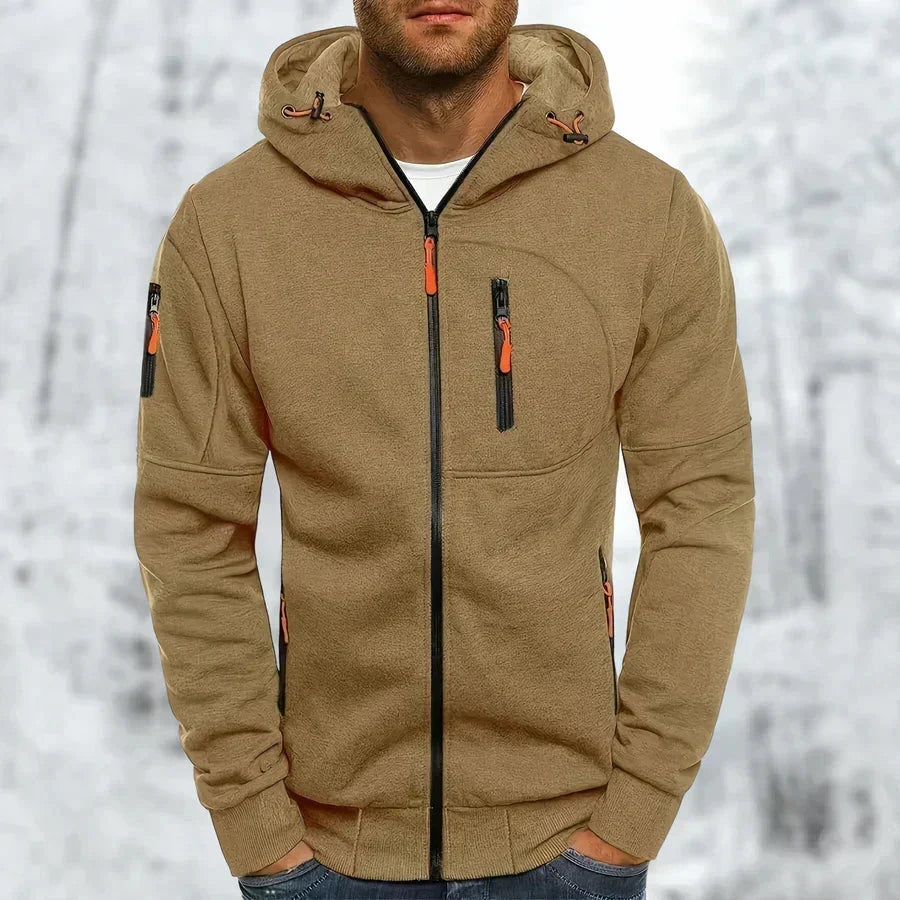 Sweat à capuche zippé beige camel porté par un modèle, zips orange et fermeture centrale noire, fond de forêt enneigée. 10
