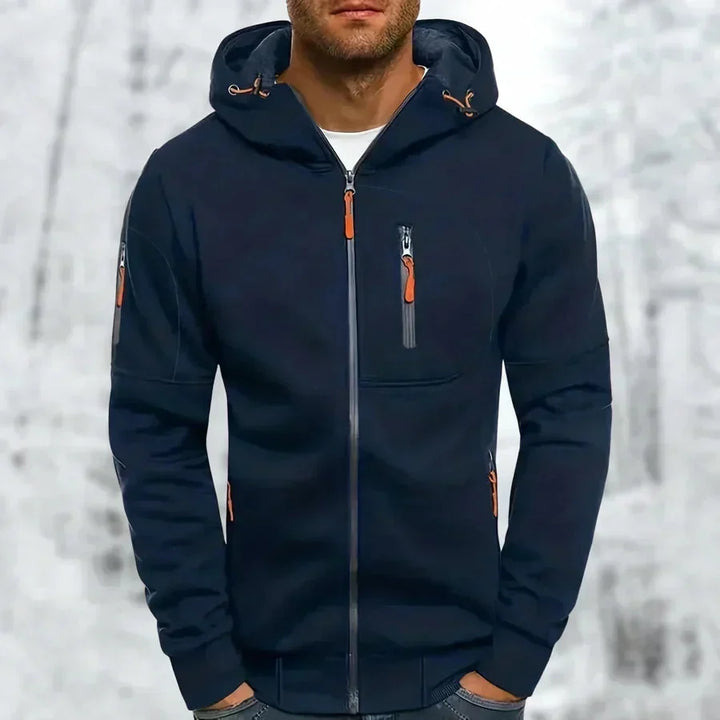 Sweat à capuche zippé bleu marine porté de face par un modèle, zips orange et poches zippées, fond enneigé. 5