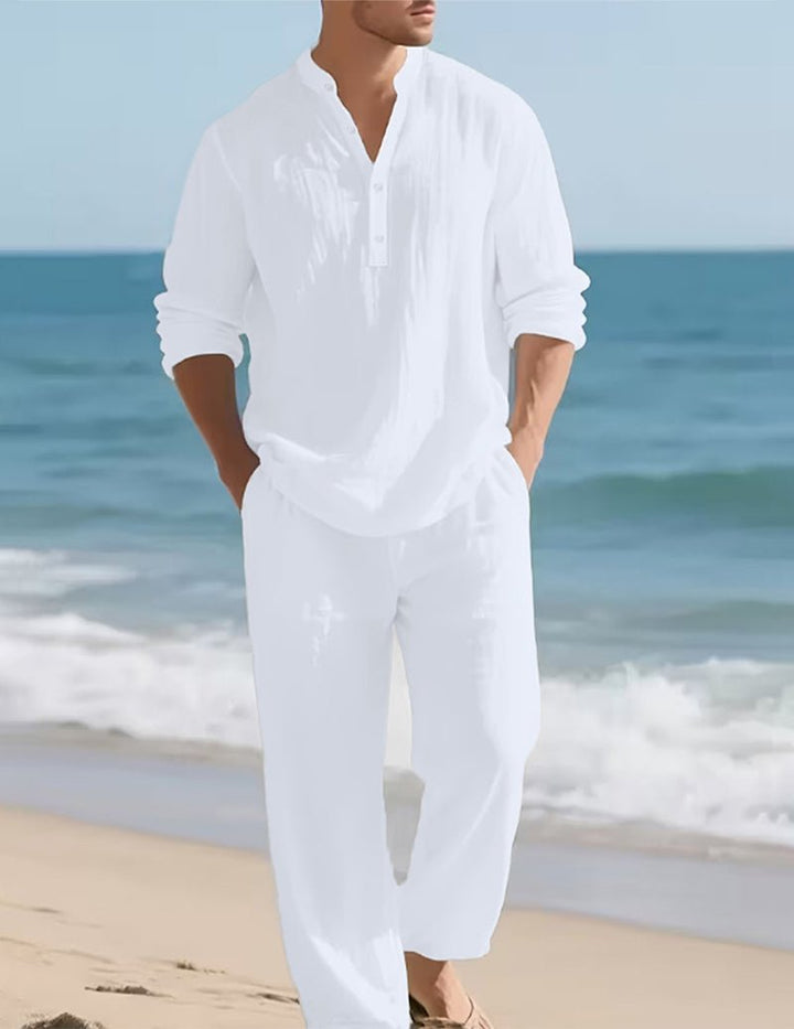 Homme en ensemble blanc marchant sur la plage au bord de l'eau, les mains dans les poches, cadré sans le visage. 2