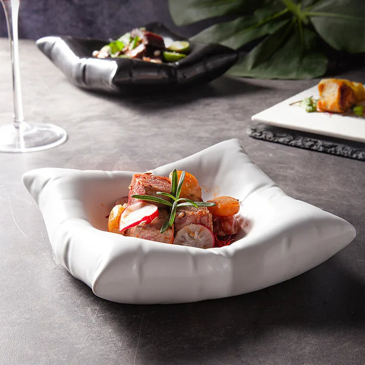 Saladier blanc aux bords plissés comme un coussin, garni de viande, radis et romarin, posé sur une table grise avec un verre et un autre plat noir. 1