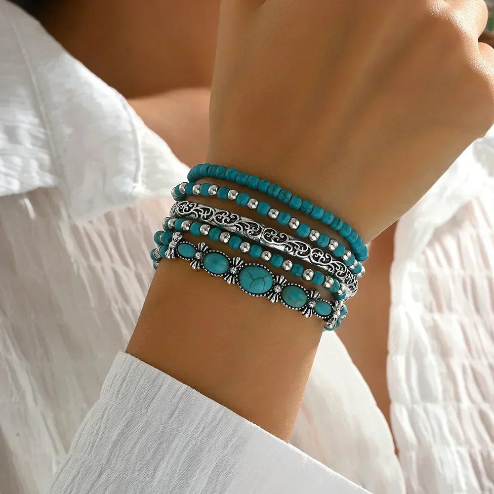 Poignet porté en gros plan avec les cinq bracelets superposés, du rang de perles au bracelet à pierres ovales, devant un chemisier blanc. 2