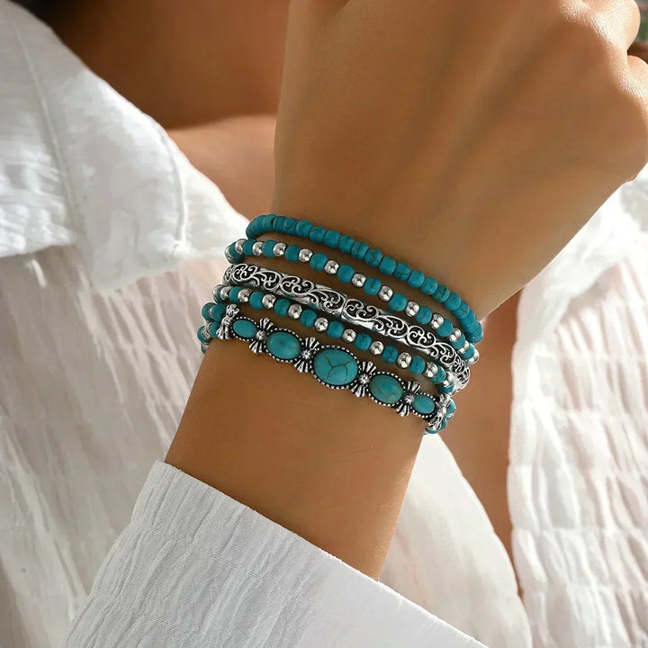 Poignet porté en gros plan avec les cinq bracelets superposés, du rang de perles au bracelet à pierres ovales, devant un chemisier blanc. 2
